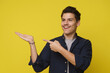20s young man with excited happy face pointing right by finger to his opened palm and looking to direction isolated over yellow studio background