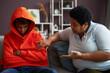 African American mature woman talking to sulky or stressed teenage grandson in red hoodie while sitting on couch next to him