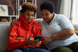 © Seventyfour - African American teenage boy in red hoodie showing online video in smartphone to his grandmother while both sitting on couch