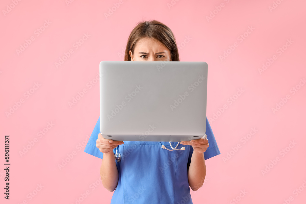 Upset female medical intern with laptop on pink background