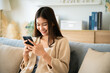 © Monster Ztudio - Happy young asian woman relax on comfortable couch at home texting messaging on smartphone, smiling girl use cellphone chatting, browse wireless internet on gadget, shopping online from home