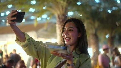 Wall Mural - Solo traveler asian women enjoy buying and eating traditional street food night market take photo upload social media. Beautiful Tourists woman feeling happy buying street food in the evening.