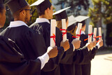 Fototapeta  - Group of diverse university students on graduation day. Happy mixed race multiethnic college graduates in black caps and gowns holding diplomas in hands while standing in row at outdoor ceremony