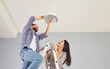 © Studio Romantic - Happy family couple changing a LED lightbulb at home. Joyful young man and woman standing on a step ladder and changing an energy-saving light bulb in a white lamp on the ceiling