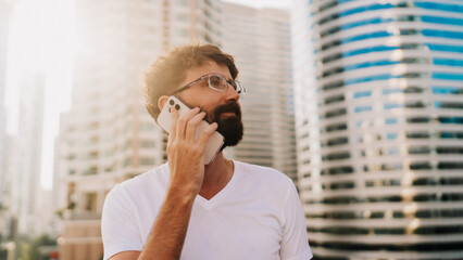 Handsome man with beard  talking by mobiyle phone while walking in big modern city with skyscrapers. Outdoors at sunset . Wearing white t shirt.