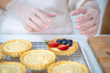 Wall Mural - close up of hands in cooking gloves Baker adding blueberries strawberry fresh fruit to a tart on white table in Kitchen. housewife baker wear apron making fruit tart. homemade bakery at home.