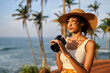 Young african female model tourist with camera in colorful clothes and straw hat taking pictures at tropical location at sunrise. Black travelling woman takes photos in exotic ocean scenery at dawn.