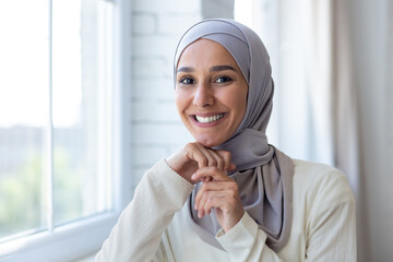 Close-up portrait of young beautiful arab woman, muslim woman in hijab smiling and looking at camera, standing near window at home.