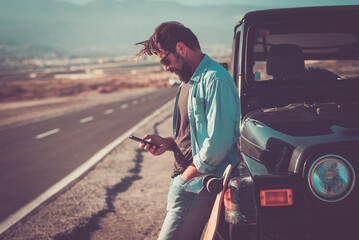 Wall Mural - One man traveler using phone and roaming connection outside the car with long scenic straight road in background. Adventure and modern lifestyle people. Adult having relax outdoor against a vehicle