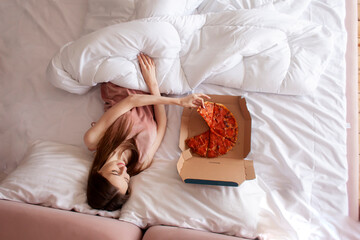 cute woman lies in bed under the covers and eats delicious pizza in the morning, woman in pajamas eats fast food