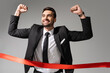 © LIGHTFIELD STUDIOS - young successful businessman crossing red finish ribbon and showing triumph gesture isolated on grey.