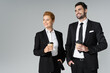 © LIGHTFIELD STUDIOS - joyful business partners holding coffee to go while standing with hands in pockets and looking away isolated on grey.