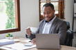 © PHAISITSAWAN - African American businessman in a black suit holding a cup of coffee to relax after checking company work on a laptop inside the office.