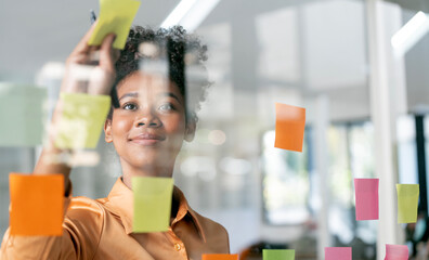 Young smiley attractive african businesswoman using sticky notes in glass wall to writing strategy business plan to development grow to success