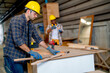 © narong - Professional Caucasian carpenter man work with woodwork in front of American African co-worker in the background of workplace.