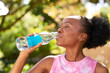 © Meeko Media - Close up of young Black woman drinking water from glass bottle in park, yoga fun
