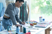 Asian LGBTQ Guy Working At Desk, Mannequin In The Background, Dressmaker, Fashion Designer Or Tailor At Work In Studio, Small Business Entrepreneur And Dressmaker Or Designer Concept