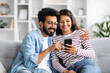 © Prostock-studio - Relaxed multicultural couple sitting on sofa at home, using smartphone