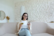 © Liubomir - A young woman is sitting on the sofa at home and using the phone. Holds a credit card and makes remote purchases, rests, has fun.