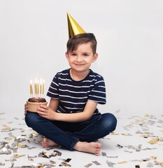 A boy of 7 years old in a striped T-shirt and jeans in a gold cap celebrates his birthday. He holds a donut with lit candles in his hands on a white background. The concept of children's birthday, chi