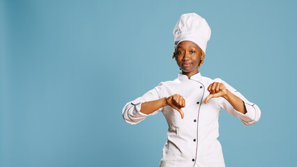 Wall Mural - Displeased female cook showing thumbs down sign, advertising disagreement and doing dislike symbol on camera. Young gourmet chef expressing refusal and rejection in studio, negative gesture.