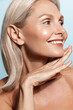 © Liubov Levytska - Smiling middle aged woman, 50 years model, applies anti-aging skin cream, rubbing in moisturizer on face, standing over blue background