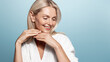 Beauty and wellbeing. Mature woman with glowing, clear skin without aging wrinkles, no blemishes, smiling coquettish, wearing spa bathrobe, doing makeup skincare procedures at home, blue background