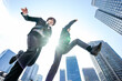 高層ビルに囲まれた都会の空をローアングルでジャンプするスーツの2人のビジネスパーソンの男性
