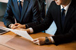 オフィスの会議テーブルで横の人物に書類の資料説明をするスーツのビジネスマンの男性の手元のパーツカット