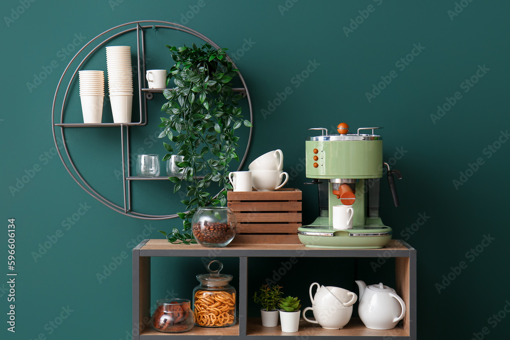 Shelving unit with modern coffee machine, cups and snacks near green wall