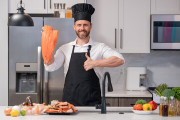 Wall Mural - Portrait of cook man preparing fresh salmon at kitchen. Handsome man in cook apron and chef hat preparing raw fish salmon. Chef cooking seafood in kitchen. Millennial man hold raw fish salmon.