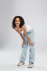 Wall Mural - full length of carefree african american woman in blue jeans smiling at camera on grey background.