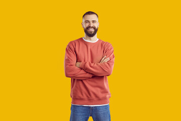 Young smiling bearded man in red sweatshirt and jeans is posing with arms crossed on chest standing isolated on yellow background looking at camera. Sincere human emotions, body language concept.