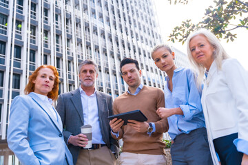 Portrait of cheerful group of coworkers laughing and looking at a tablet outdoors in a corporate office area, businesswoman and businessman and executives in commercial area