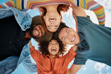 Canvas Print - Group of diverse friends standing in a huddle against a blue sky. Multi-racial friends standing together arms around each other and smiling while looking down at the camera