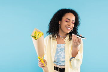 Wall Mural - positive african american woman with yellow tulips sending voice message on smartphone isolated on blue.