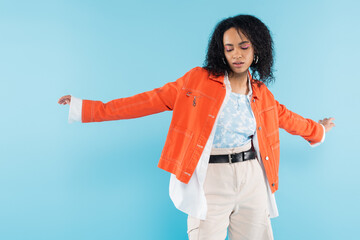 Wall Mural - trendy african american woman in orange jacket and shorts posing with outstretched hands isolated on blue.