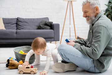 bearded man smiling near toddler granddaughter playing with toy car near building blocks on floor in living room.