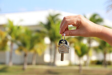 Real Estate Agent, Home Keys In Female Hand On Background Of House Surrounded By Palm Trees. Buying Or Rental A Villa On Ocean Coast, Holidays Or Removal To Tropical Country