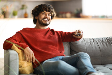 Wall Mural - Weekend Pastime. Cheerful Indian Guy Watching Tv At Home, Holding Remote Controller