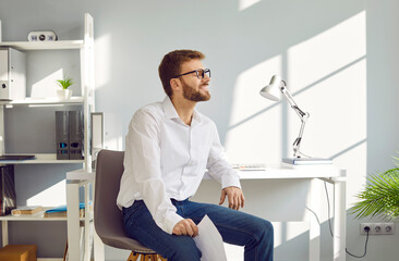 Portrait of a joyful young happy attractive bearded business man in glasses and white shirt sitting at his workplace in the office with closed eyes having rest from a work, smiling and relaxing.