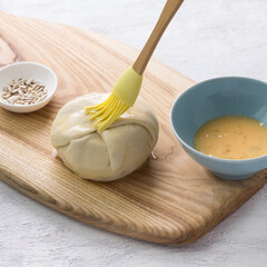 Small raw pie smeared with egg before baking surrounded by two bowls with a beaten egg and seeds on light gray background. Stage of cooking delicious homemade food