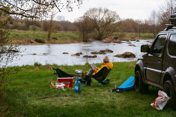 a woman is reading a book. an off-road vehicle with a roof rack. camping equipment. view on river. c