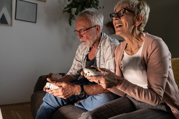 Senior couple sitting on sofa and playing video game on console. They're holding a game pad and challenge each other to win.	
