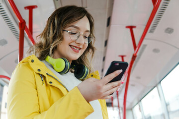 Portrait of young smiling woman passenger with headphones using smartphone in the bus.