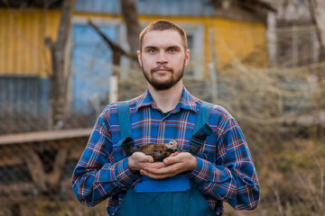 A satisfied man farmer of European Caucasian appearance, bearded, in a shirt and overalls, holds a dwarf chicken in his hands against the background of the countryside