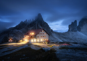 Wall Mural - mountains in fog with beautiful house with lights at night in summer. landscape with high rocks, blu