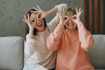 Close up two adult women mature mom young kid wear casual clothes hold hand near eyes pov glasses hug sit on gray sofa couch stay at home flat rest relax spend free time in living room Family concept