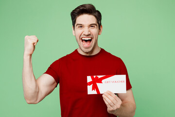 Wall Mural - Young surprised happy man he wearing red t-shirt casual clothes hold gift certificate coupon voucher card for store do winner gesture isolated on plain pastel light green background studio portrait.