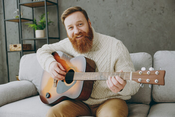 Wall Mural - Young smiling fun cheerful ginger man wear casual clothes play guitar look camera sit on grey sofa couch stay at home hotel flat rest relax spend free spare time in living room indoor. Lounge concept.